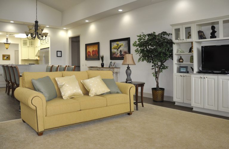 A spacious living room with a yellow sofa, white built-in shelving, television, and open kitchen area.